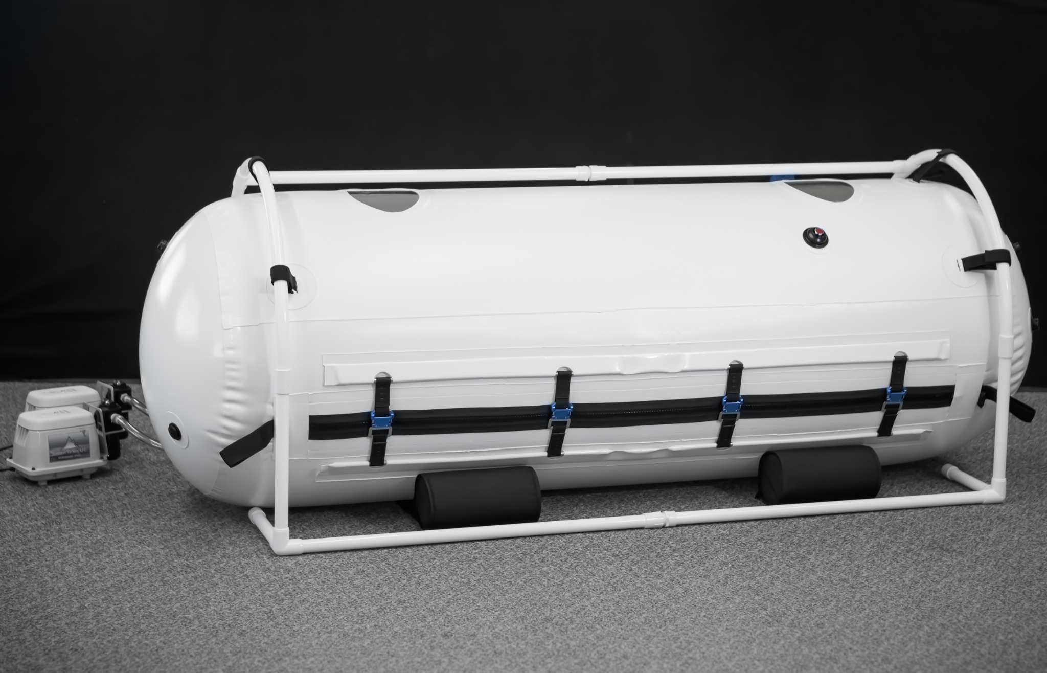 Biała, przenośna komora hiperbaryczna na białym stelażu, z widocznymi czarnymi pasami i niebieskimi klamrami, oraz dwoma pompami powietrza z boku, na szarym dywanie i czarnym tle.