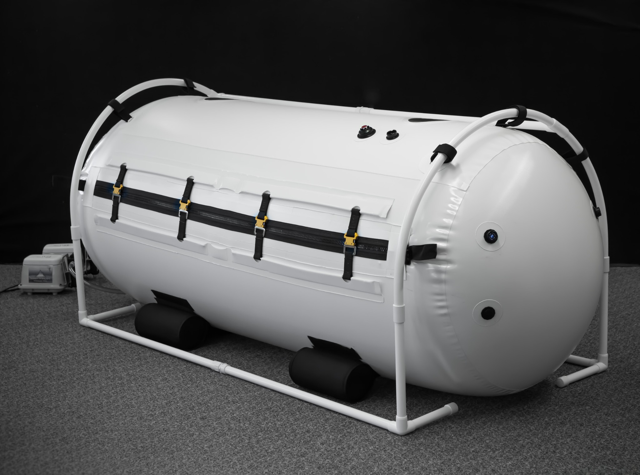 Biała, przenośna komora hiperbaryczna z czarnymi zamkami błyskawicznymi i złotymi klamrami, umieszczona na białej ramie z PVC, na szarym dywanie, z czarnym tłem.