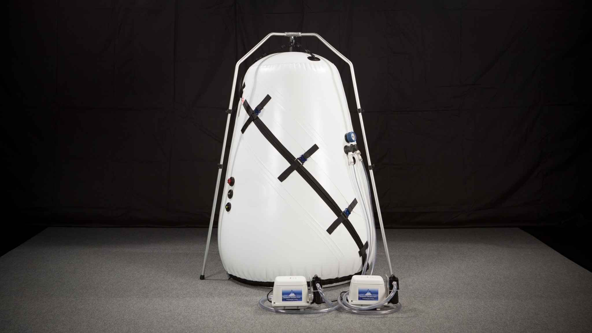 Biała komora hiperbaryczna w metalowym stelażu, z dwoma pompami powietrza na szarym dywanie i czarnym tle, widoczne złącza i węże.