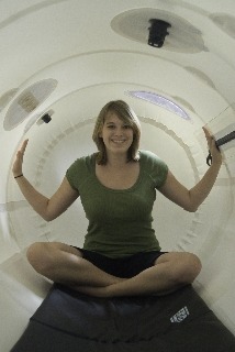 Uśmiechnięta kobieta siedząca w pozycji lotosu wewnątrz białej komory hiperbarycznej, ręce uniesione w geście powitania, widoczne okna i oświetlenie wewnątrz komory.