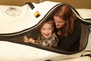 Uśmiechnięty chłopiec w komorze hiperbarycznej, nad nim uśmiechnięta kobieta, widoczne logo na komorze.