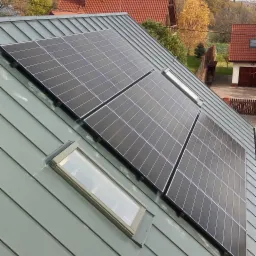 Panele słoneczne zamontowane na blaszanym dachu domu jednorodzinnego, widoczne okna dachowe i fragmenty okolicznych budynków w tle.