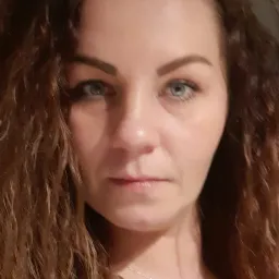 Portret kobiety z długimi kręconymi włosami i niebieskimi oczami, patrzącej prosto w obiektyw, z delikatnym srebrnym naszyjnikiem z zawieszką w kształcie kwiatu.