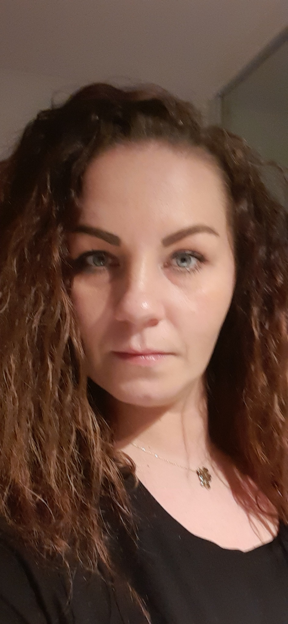 Portret kobiety z długimi kręconymi włosami i niebieskimi oczami, patrzącej prosto w obiektyw, z delikatnym srebrnym naszyjnikiem z zawieszką w kształcie kwiatu.