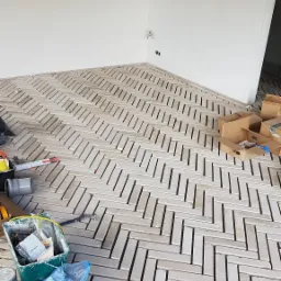 Układanie podłogi z cegiełek w jodełkę, widoczne narzędzia i materiały budowlane, surowe ściany w tle.