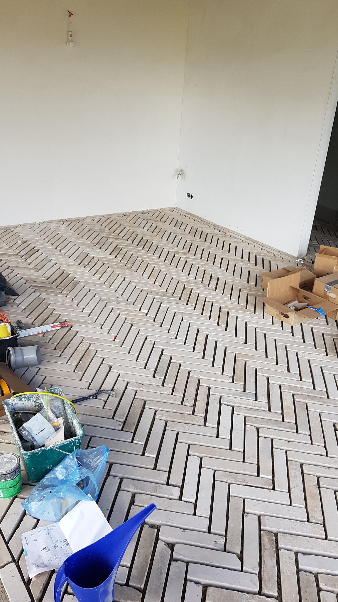 Układanie podłogi z cegiełek w jodełkę, widoczne narzędzia i materiały budowlane, surowe ściany w tle.