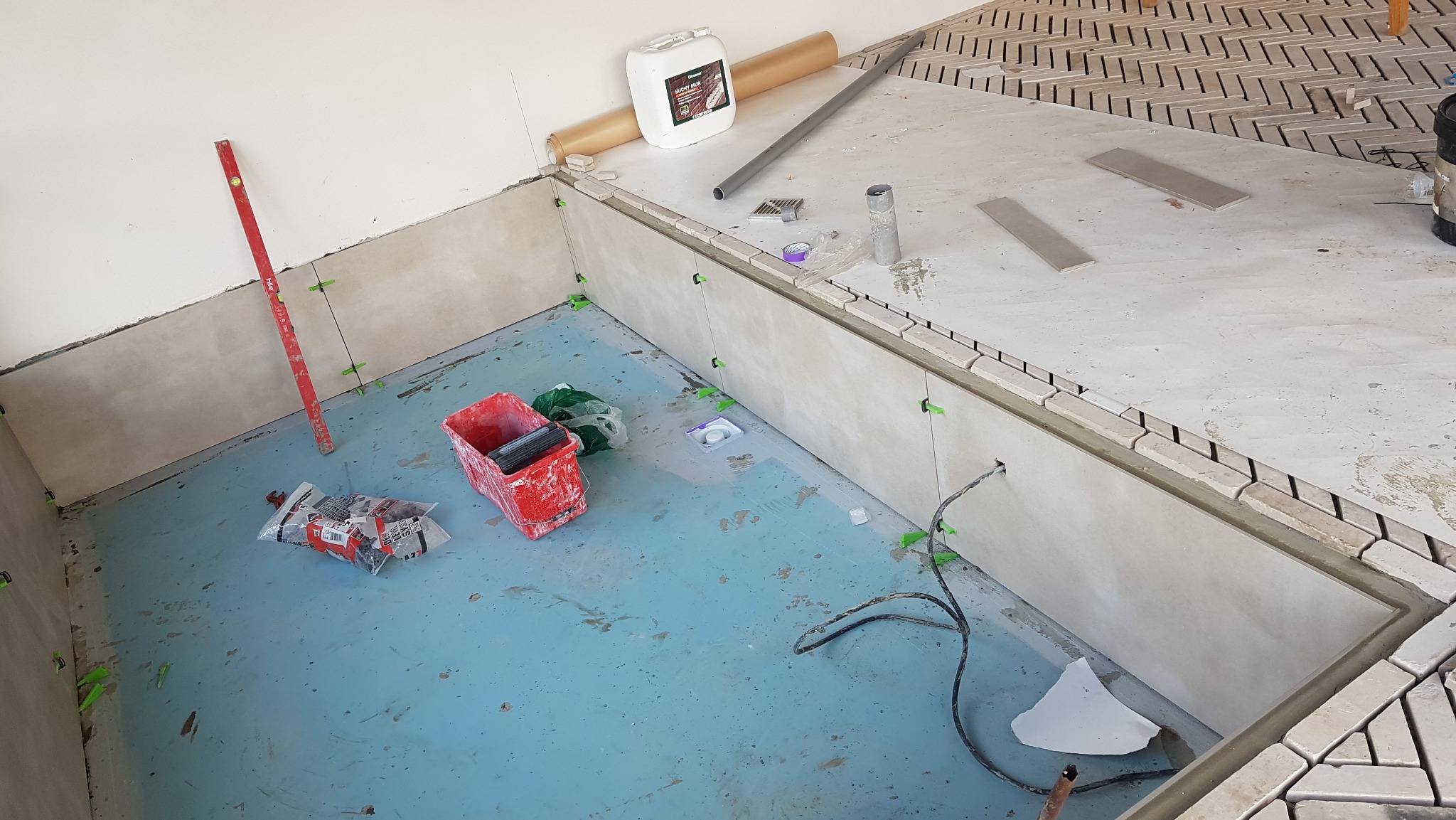 Etap prac glazurniczych: układanie mozaiki w kształcie jodełki na podłodze i ścianach, widoczne poziomica, pojemnik z płytkami, opakowania po kleju i zielone kliny dystansowe.