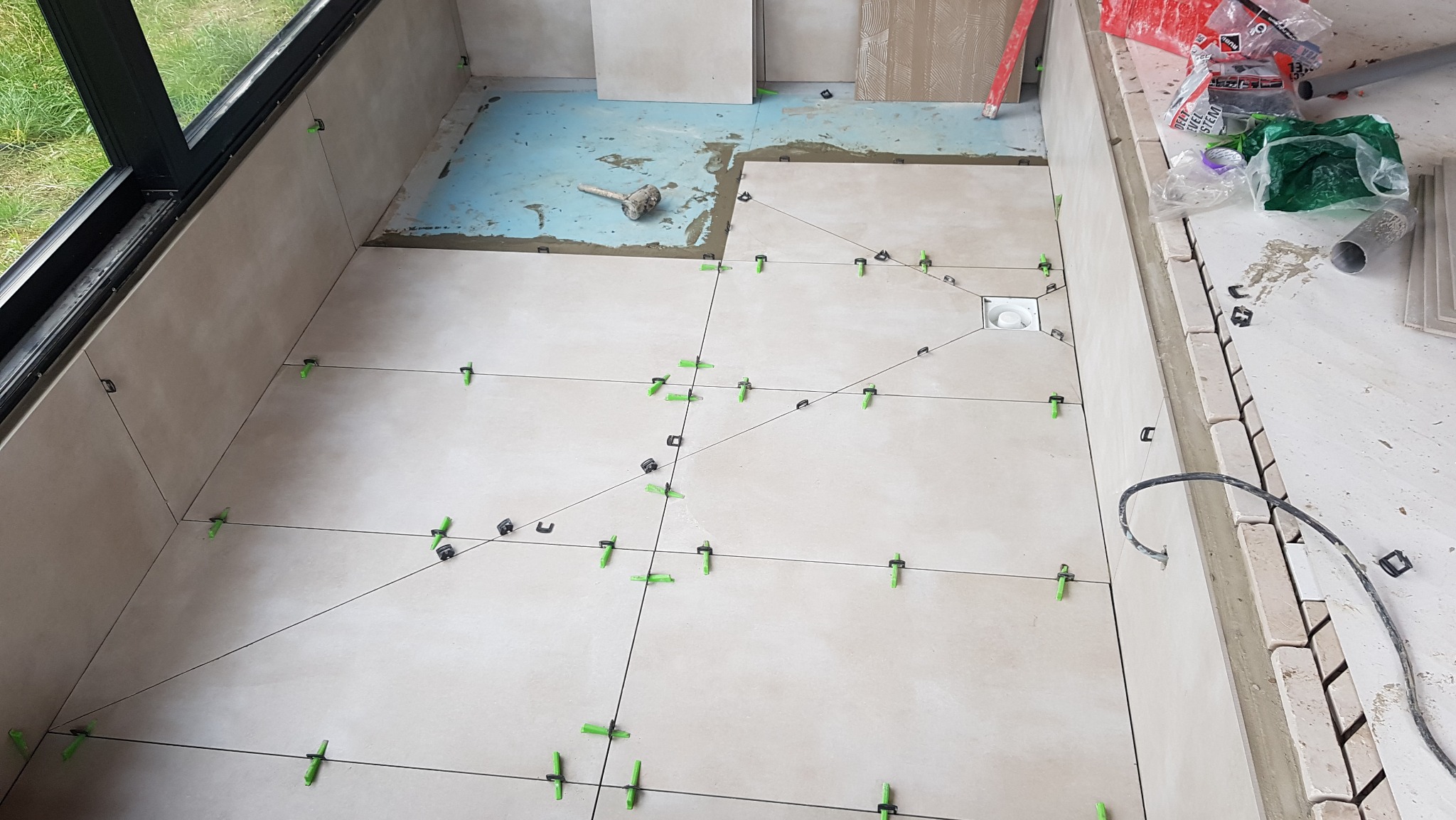 Układanie beżowych płytek podłogowych z użyciem systemu poziomowania, widoczny odpływ liniowy, fragment okna i narzędzia pracy.