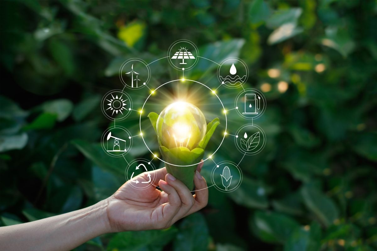 Żarówka LED otoczona liśćmi, symbolizująca ekologię, z nałożonymi ikonami różnych źródeł energii odnawialnej na tle zieleni.