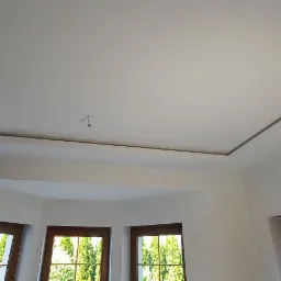 Nowoczesny sufit podwieszany z podświetleniem obwodowym w jasnym pomieszczeniu z oknami i drewnianą stolarką.