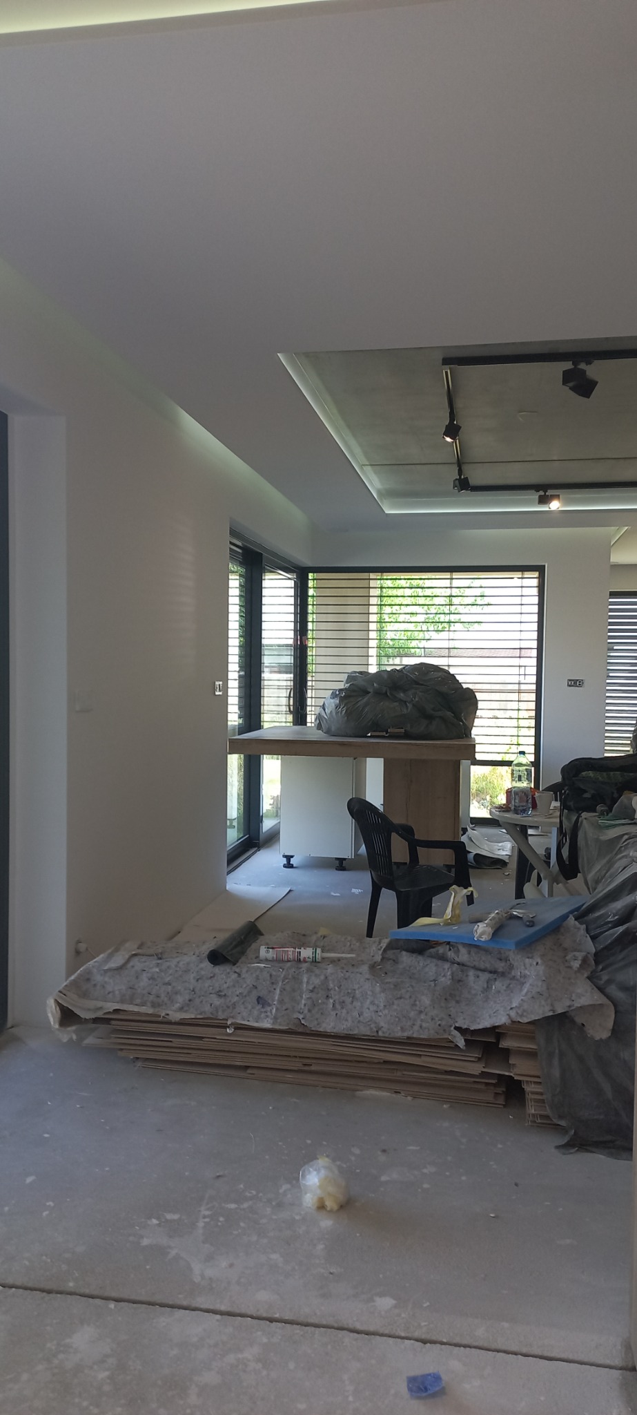 Wnętrze w trakcie prac wykończeniowych: widoczne płyty, narzędzia i worki z materiałami budowlanymi na podłodze, świeżo pomalowane ściany i sufit z oświetleniem punktowym.