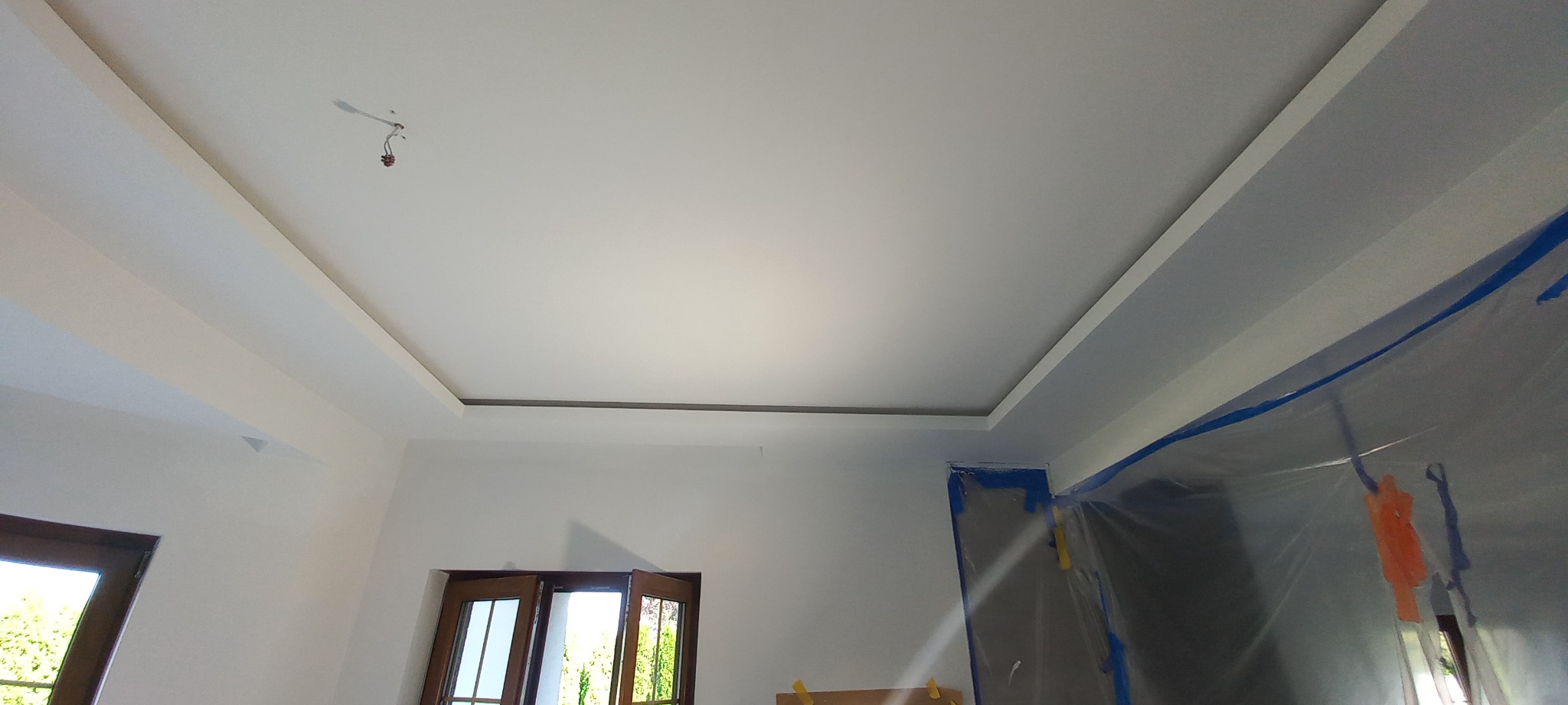 Wnętrze po remoncie: białe ściany i sufit z dekoracyjnym obniżeniem, zabezpieczona folią i taśmą malarską framuga drzwi
