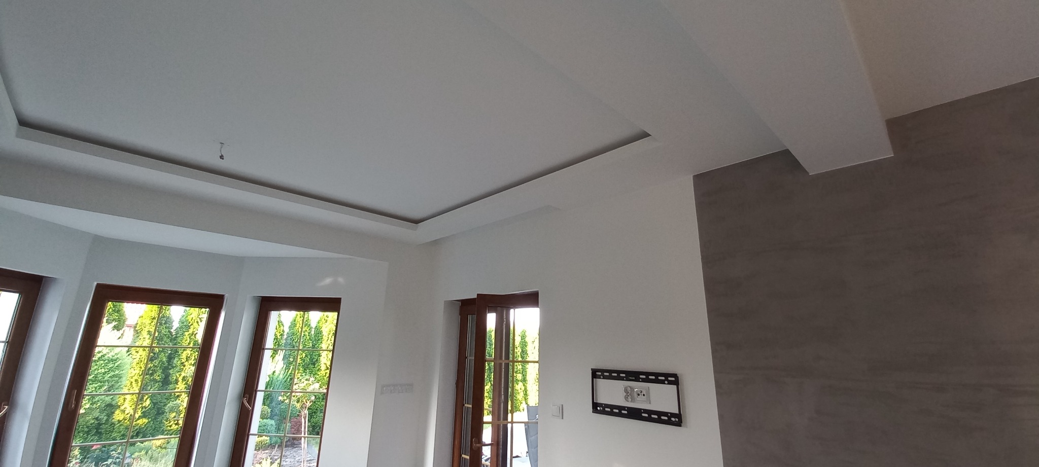 Nowoczesny salon po remoncie: białe ściany, ozdobny sufit podwieszany z oświetleniem, ściana pokryta szarą dekoracyjną farbą, widok na zieleń przez okna z drewnianą stolarką.