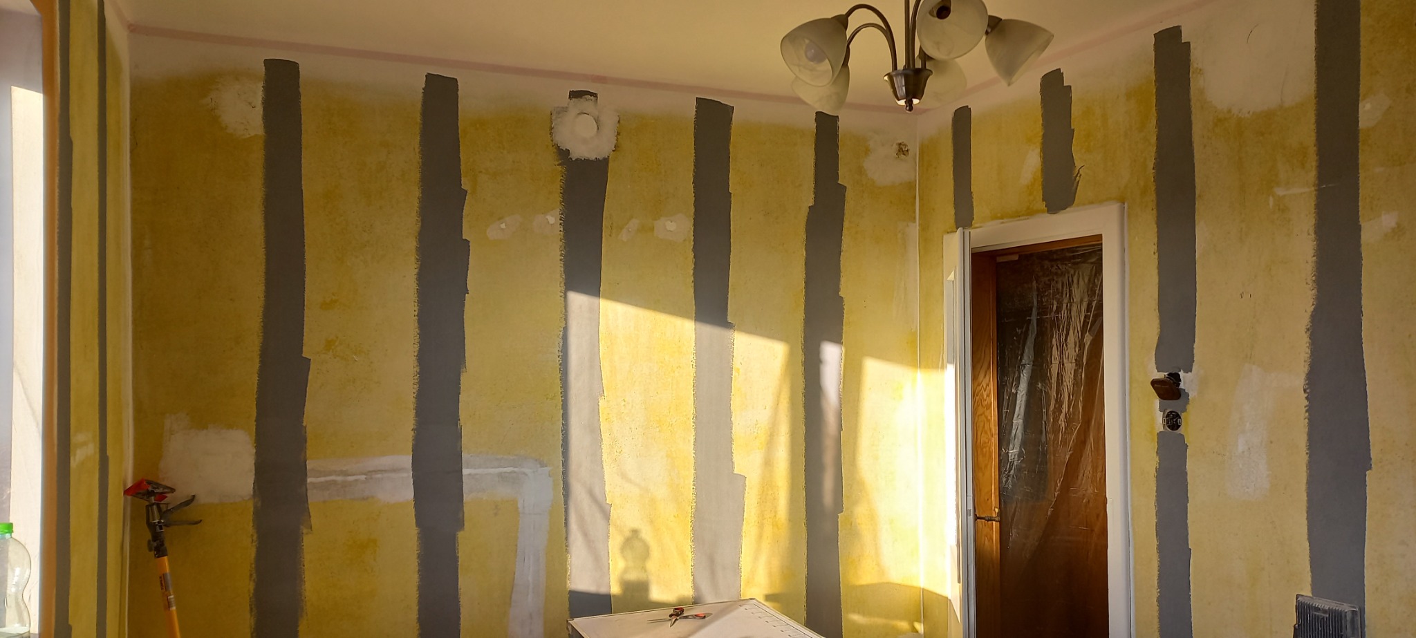 Pokój w trakcie przygotowań do malowania: ściany z żółtą tapetą, pionowe pasy szarej farby gruntującej, widoczne punkty naprawcze i zakryte drzwi folią ochronną.