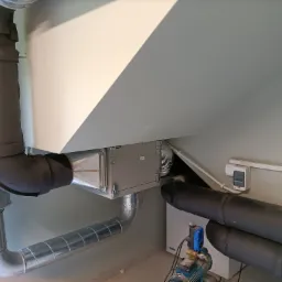 Centralna jednostka wentylacyjna z kanałami, widoczne rury izolowane termicznie oraz pompa ciśnieniowa, całość zainstalowana w pomieszczeniu ze skośnym sufitem.