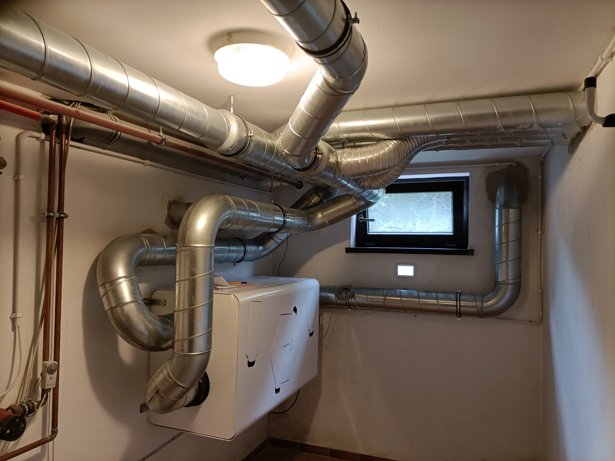 Instalacja rekuperacji z białym rekuperatorem i systemem metalowych rur wentylacyjnych pod sufitem w pomieszczeniu z oknem.