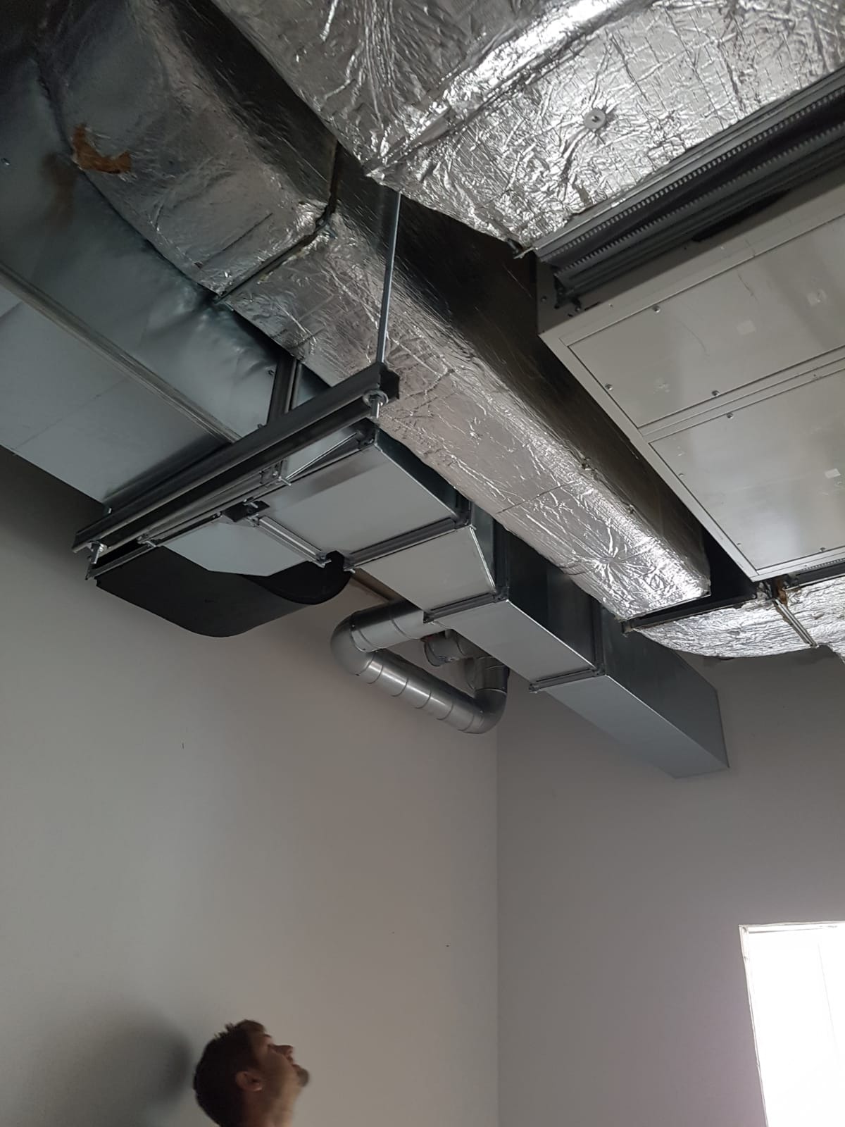 System kanałów wentylacyjnych z izolacją termiczną, widoczny pod sufitem w pomieszczeniu, z fragmentem twarzy mężczyzny spoglądającego w górę.