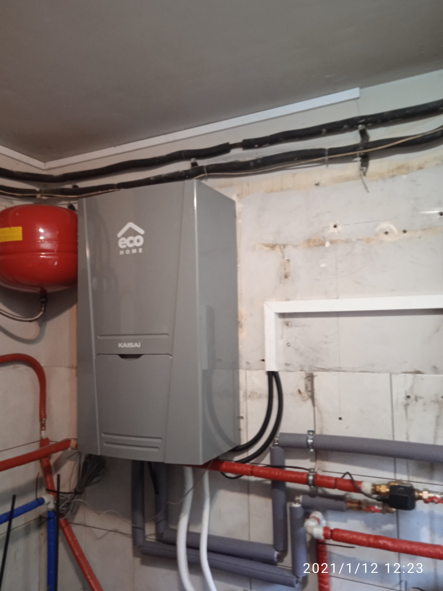 Szary moduł Kaisa ECO HOME z podłączonymi rurami wodnymi w pomieszczeniu z nieotynkowanymi ścianami i instalacją elektryczną na suficie, widoczny czerwony zbiornik wyrównawczy.