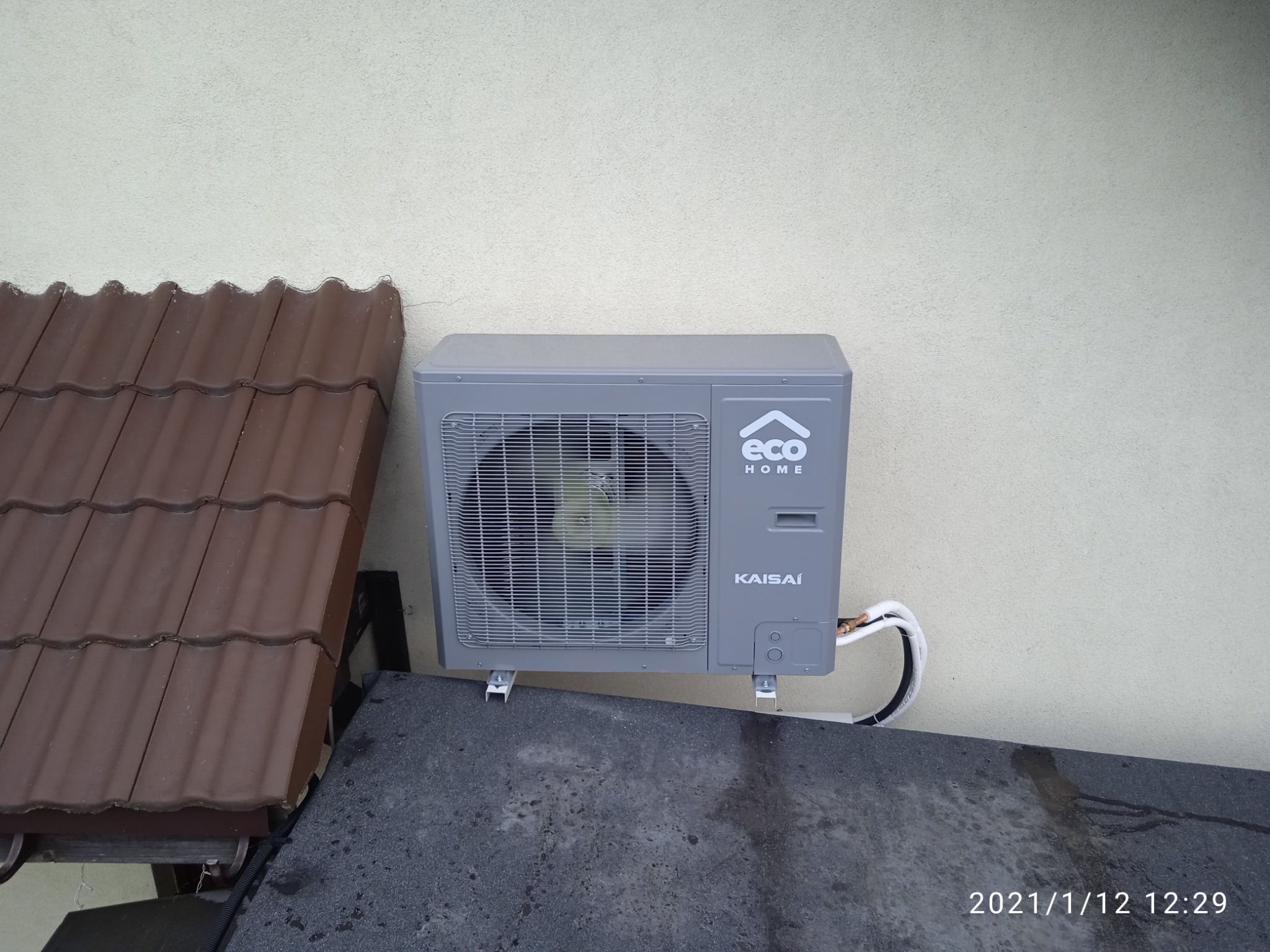 Szara jednostka zewnętrzna klimatyzatora Kaisai Eco Home zamontowana na wspornikach przy ścianie, widok z dołu, obok fragment dachu z brązowej dachówki.