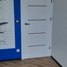 Rafał Dominski - Białe drzwi z czarnymi, poziomymi paskami na tle niebieskiej ściany z rysunkiem i drewnianej podłogi. Minimalistyczny design.