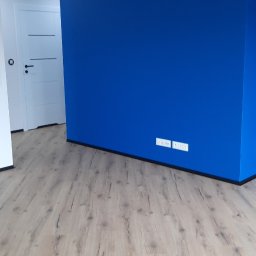 Rafał Dominski - Pokój po remoncie: świeżo pomalowana na niebiesko ściana, jasne panele podłogowe z wyraźną strukturą drewna, białe drzwi z poziomymi frezami.
