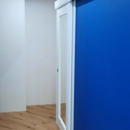 Rafał Dominski - Błękitna ściana z przesuwnymi, białymi drzwiami. Na podłodze panele imitujące drewno. W tle biała ściana i sufit z dwoma punktowymi lampami.