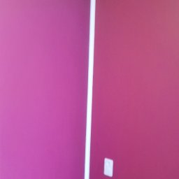 Rafał Dominski - Pomieszczenie po malowaniu ścian na dwa odcienie różu, z białymi listwami narożnymi i przypodłogowymi. Widoczny fragment drewnianej podłogi i gniazdko elektryczne.