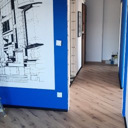 Rafał Dominski - Wykończone wnętrze: świeżo ułożone panele podłogowe, pomalowana na niebiesko ściana z grafiką i widok na korytarz. Widoczny fragment drabiny.