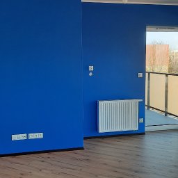 Rafał Dominski - Pomalowane na intensywny niebieski kolor ściany w pokoju z drewnianą podłogą, białym grzejnikiem i wyjściem na balkon. Widoczne gniazdka elektryczne.