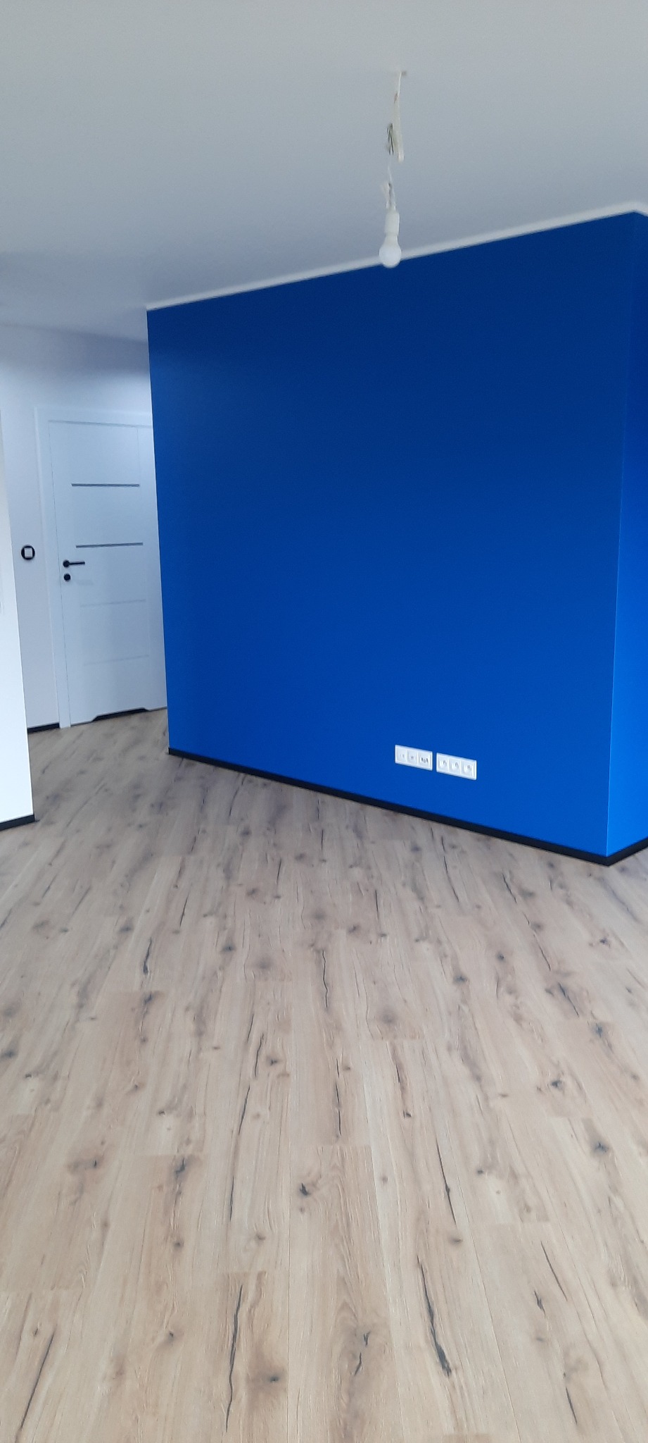 Pokój po remoncie: świeżo pomalowana na niebiesko ściana, jasne panele podłogowe z wyraźną strukturą drewna, białe drzwi z poziomymi frezami.