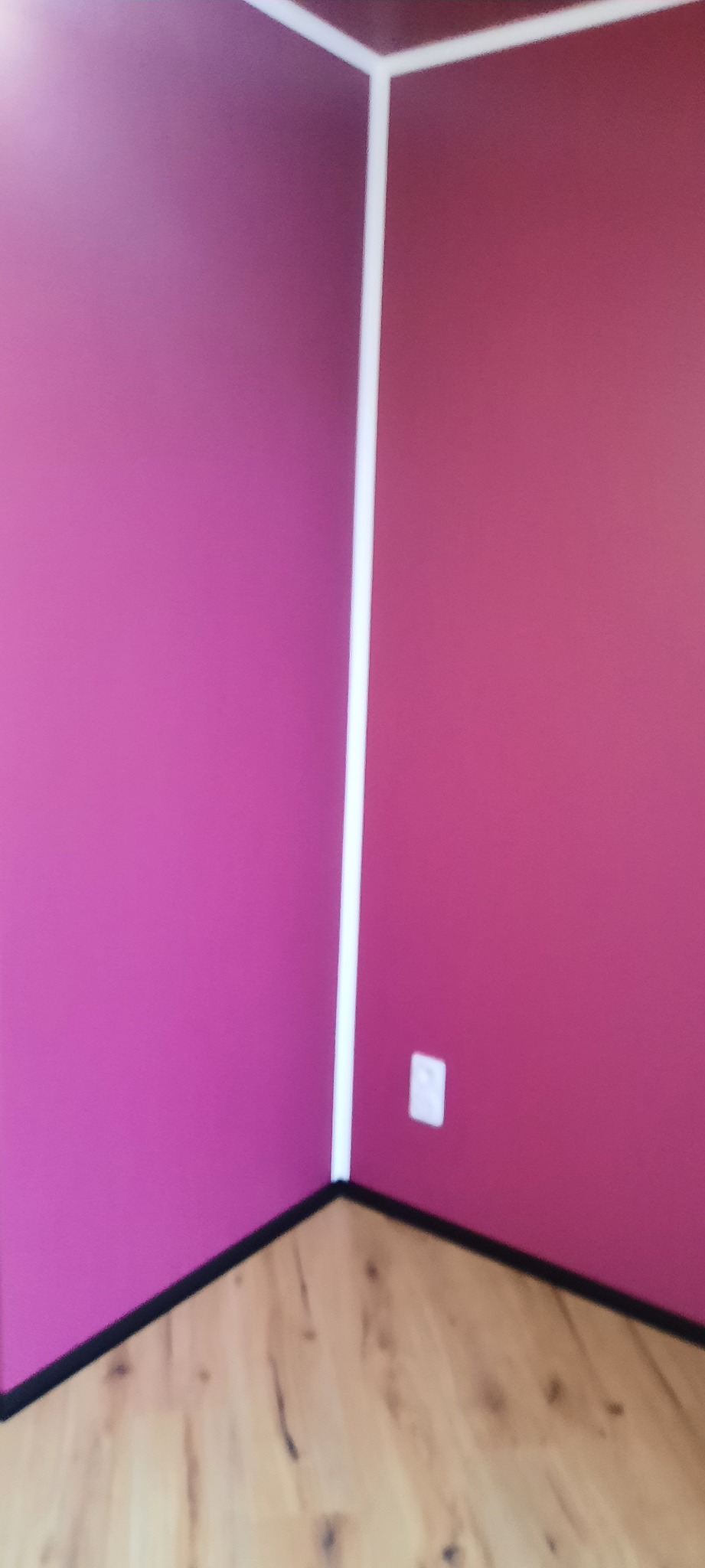 Pomieszczenie po malowaniu ścian na dwa odcienie różu, z białymi listwami narożnymi i przypodłogowymi. Widoczny fragment drewnianej podłogi i gniazdko elektryczne.