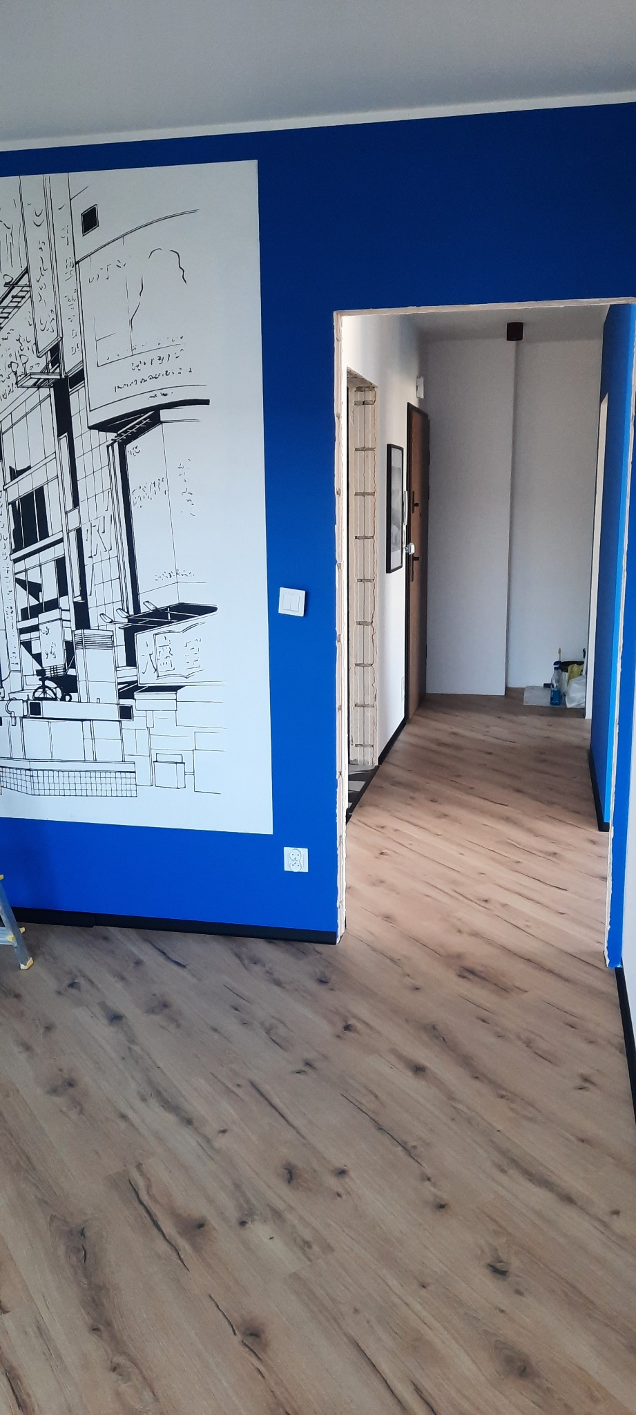 Wykończone wnętrze: świeżo ułożone panele podłogowe, pomalowana na niebiesko ściana z grafiką i widok na korytarz. Widoczny fragment drabiny.