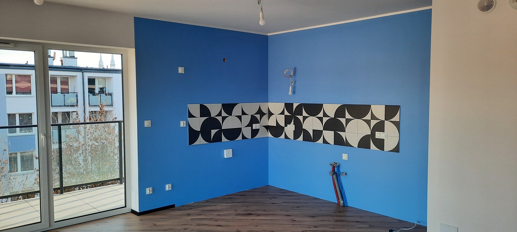 Niebieska ściana z dekoracyjną mozaiką nad blatem, widoczne instalacje i panele podłogowe. Nowoczesne wykończenie wnętrza kuchni w mieszkaniu.