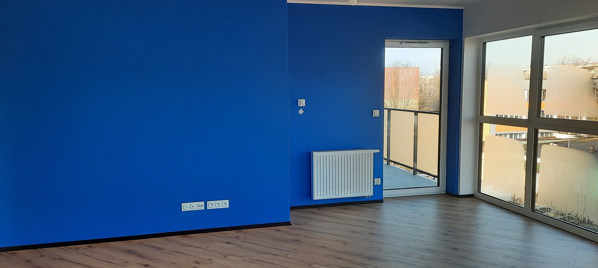 Pomalowane na intensywny niebieski kolor ściany w pokoju z drewnianą podłogą, białym grzejnikiem i wyjściem na balkon. Widoczne gniazdka elektryczne.