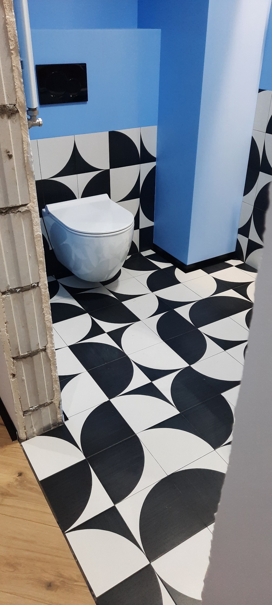 Nowoczesna łazienka z białą toaletą i geometrycznymi czarno-białymi płytkami na podłodze i ścianach. Niebieski element zabudowy. Widoczny fragment surowej ściany.