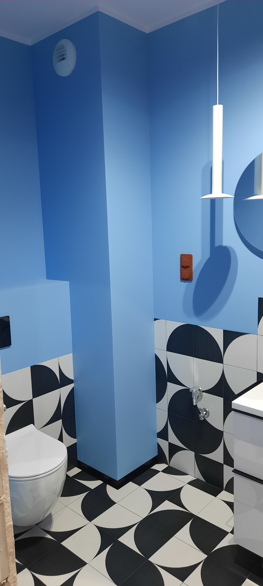 Niebieska łazienka z geometrycznymi płytkami: toaleta, umywalka i kolumna w odcieniach błękitu. Nowoczesne oświetlenie i brązowy włącznik dodają charakteru.