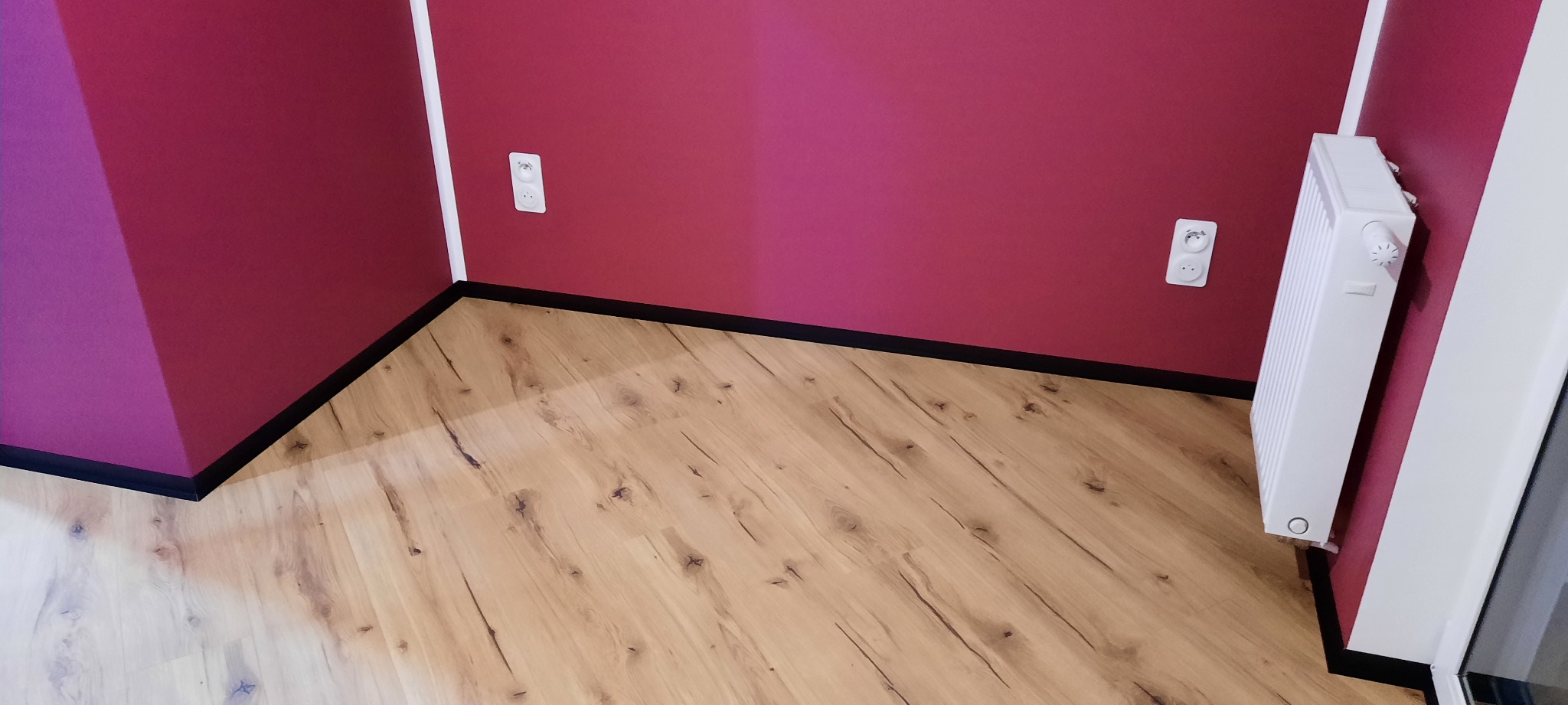 Wnętrze po remoncie: panele podłogowe w odcieniach drewna, ściany pomalowane na intensywny odcień czerwieni, biały grzejnik i gniazdka elektryczne.