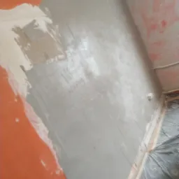 Ściana w trakcie prac przygotowawczych, częściowo pokryta warstwą szarej masy szpachlowej, z widocznymi fragmentami starej farby w kolorze pomarańczowym i białym. Podłoga zabezpieczona folią.