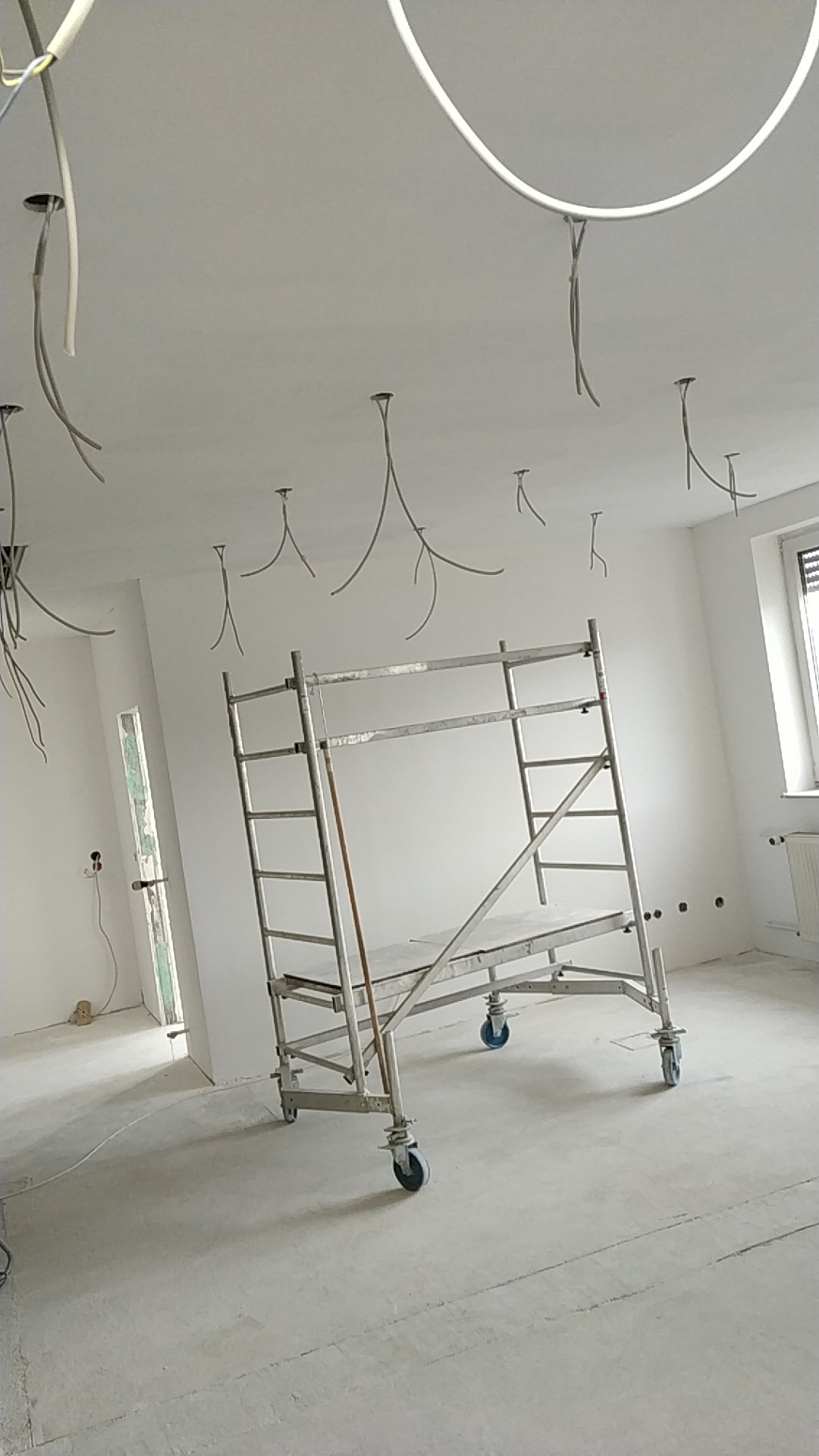 Wnętrze w trakcie prac wykończeniowych; widoczne kable zwisające z sufitu, aluminiowe rusztowanie na kółkach, ściany przygotowane do malowania lub tapetowania, surowa podłoga.