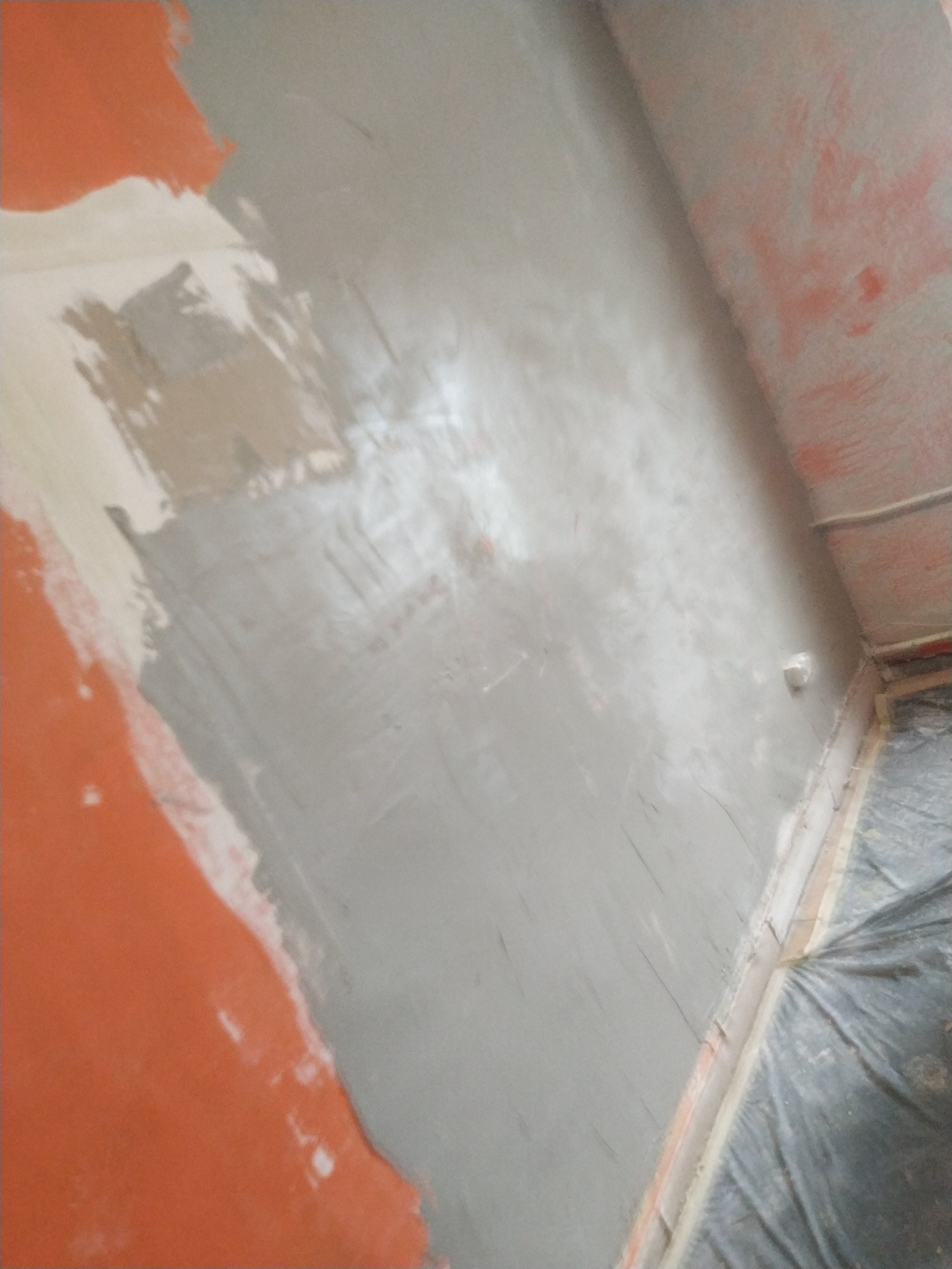 Ściana w trakcie prac przygotowawczych, częściowo pokryta warstwą szarej masy szpachlowej, z widocznymi fragmentami starej farby w kolorze pomarańczowym i białym. Podłoga zabezpieczona folią.