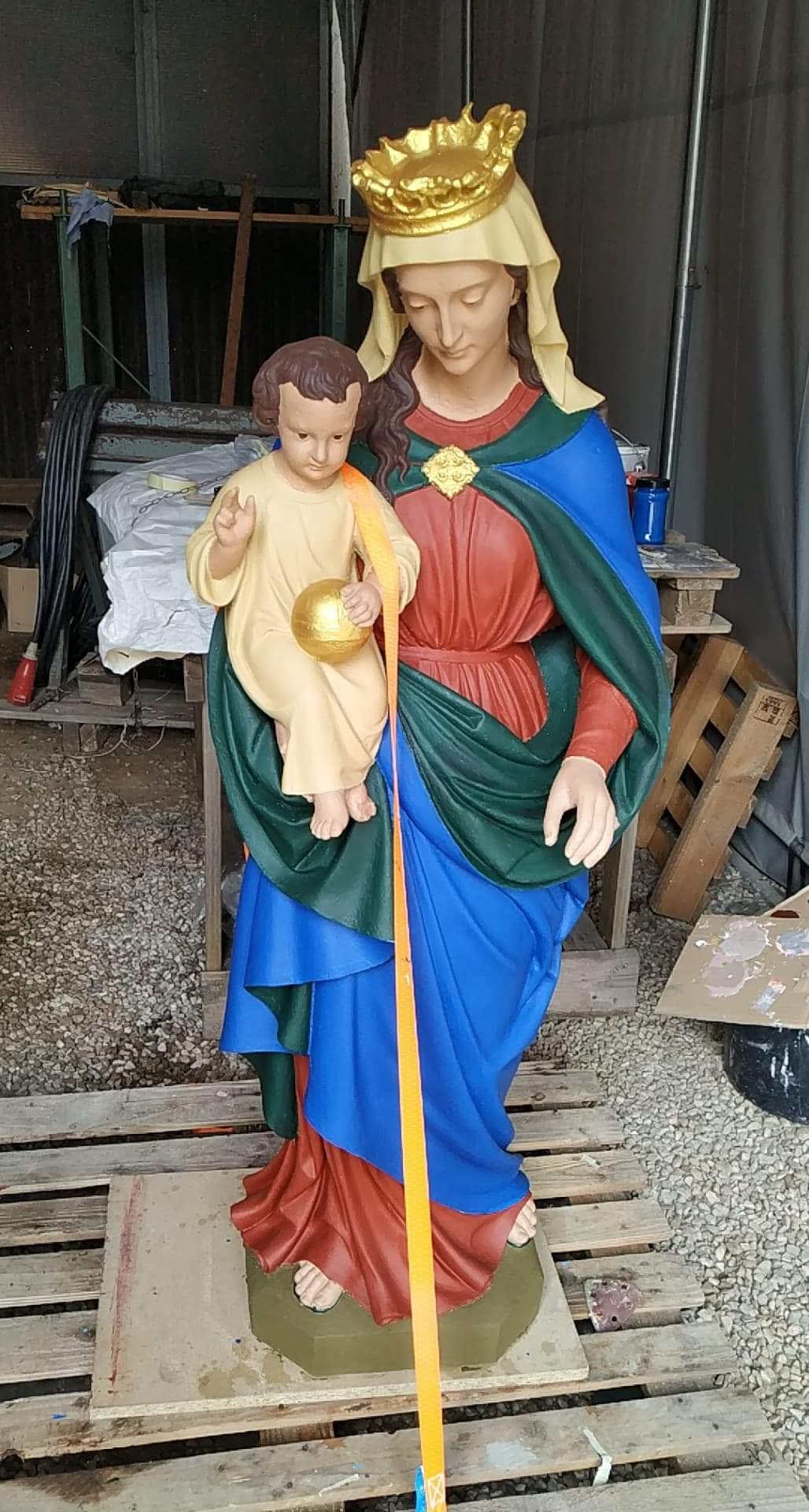 Figurka Matki Boskiej z Dzieciątkiem po renowacji, z widocznymi świeżymi warstwami farby i paskiem zabezpieczającym, umieszczona na palecie w warsztacie.