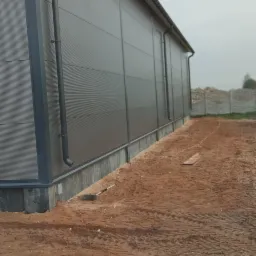 Szara hala z płyt warstwowych z widocznym systemem rynnowym, położona na gruncie z piasku, z widocznymi śladami opon i narzędziem budowlanym leżącym obok ściany.