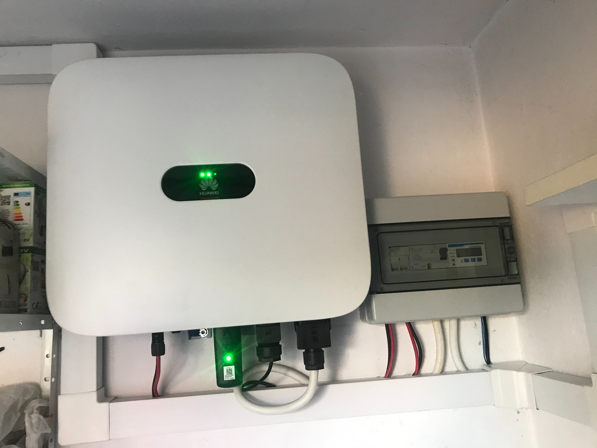 Biały falownik Huawei z zielonymi diodami LED i podłączonymi kablami, obok skrzynka z bezpiecznikami na białej ścianie.