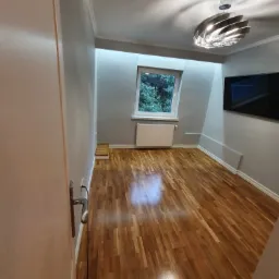 Pomalowane na szaro wnętrze pokoju z drewnianą podłogą, oświetlone nowoczesną lampą sufitową, z widocznym oknem i grzejnikiem.