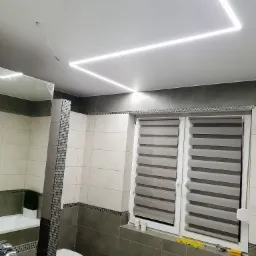 Nowoczesna łazienka z kwadratowym oświetleniem LED na suficie, szarymi płytkami i roletami okiennymi w paski. Widoczny fragment wanny, toaleta oraz umywalka z lustrem.