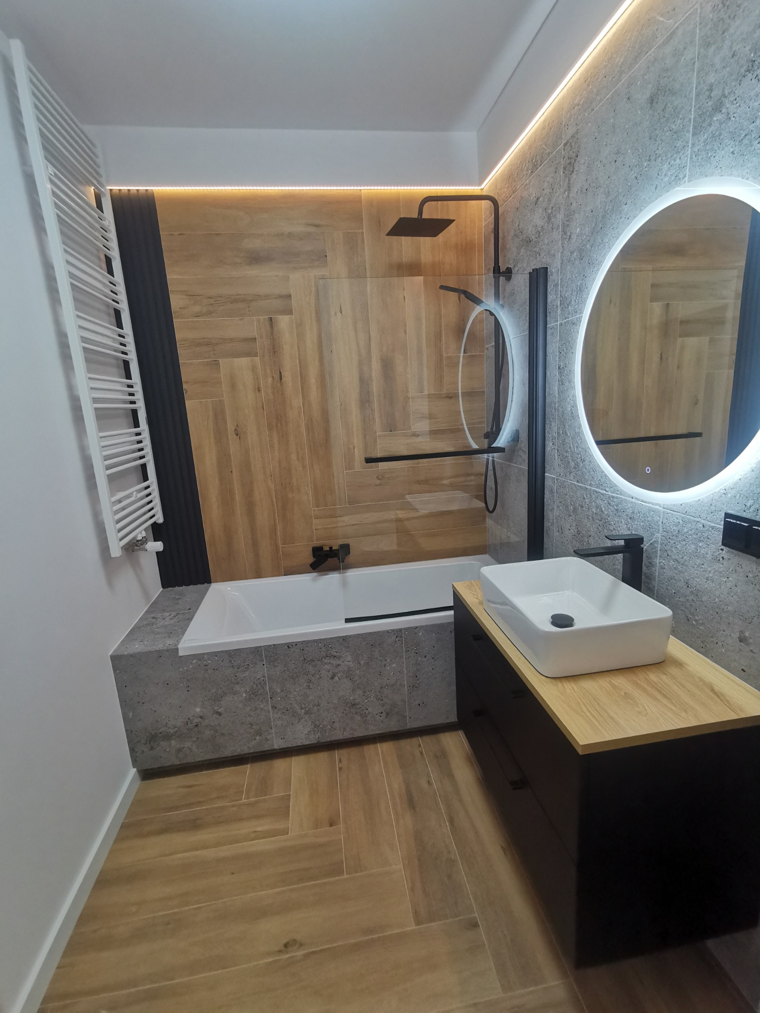 Nowoczesna łazienka z drewnopodobnymi płytkami ułożonymi w jodełkę, czarną armaturą, okrągłym lustrem z podświetleniem LED i białą, prostokątną umywalką nablatową.
