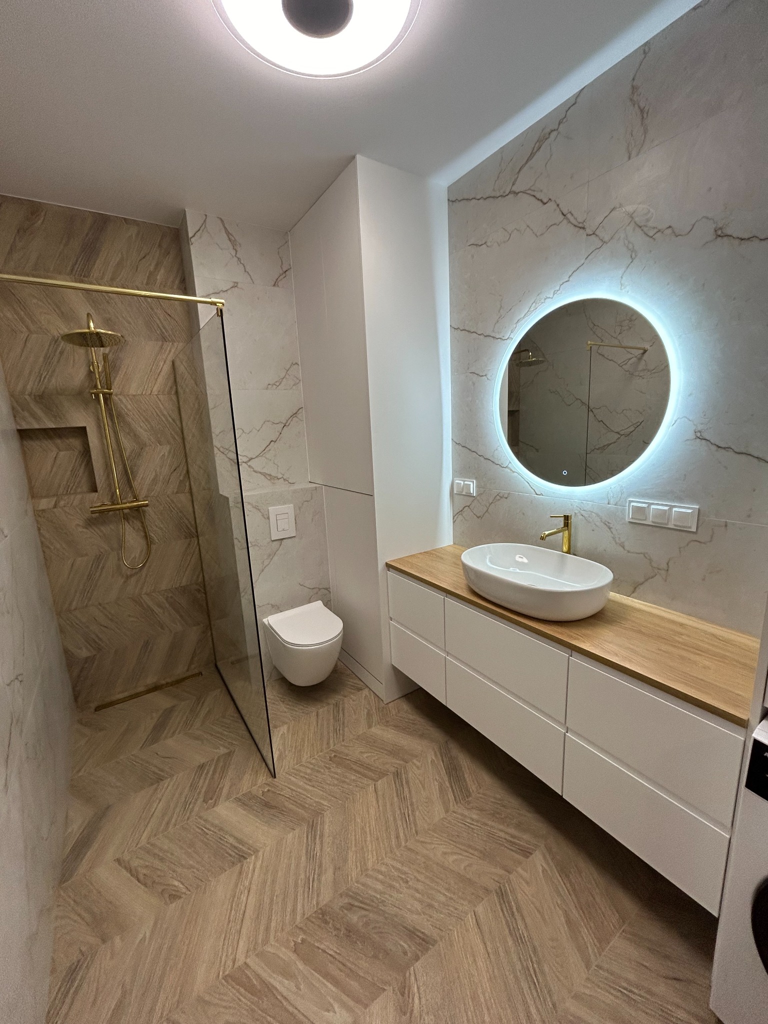 Nowoczesna łazienka z kabiną prysznicową typu walk-in z złotą armaturą, umywalką nablatową na drewnianym blacie i okrągłym lustrem z podświetleniem LED, wykończona płytkami imitującymi marmur.