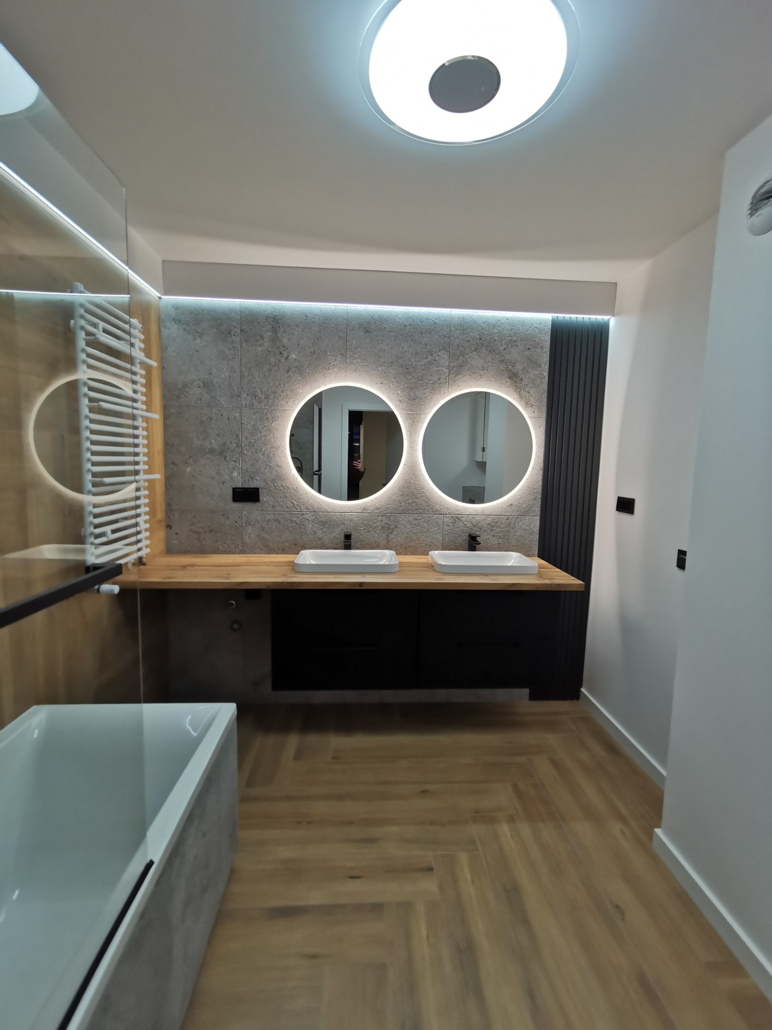 Nowoczesna łazienka z drewnianą podłogą w jodełkę, dwoma okrągłymi lustrami z podświetleniem LED nad dwiema umywalkami nablatowymi, drewnianym blatem, grzejnikiem drabinkowym i wanną.