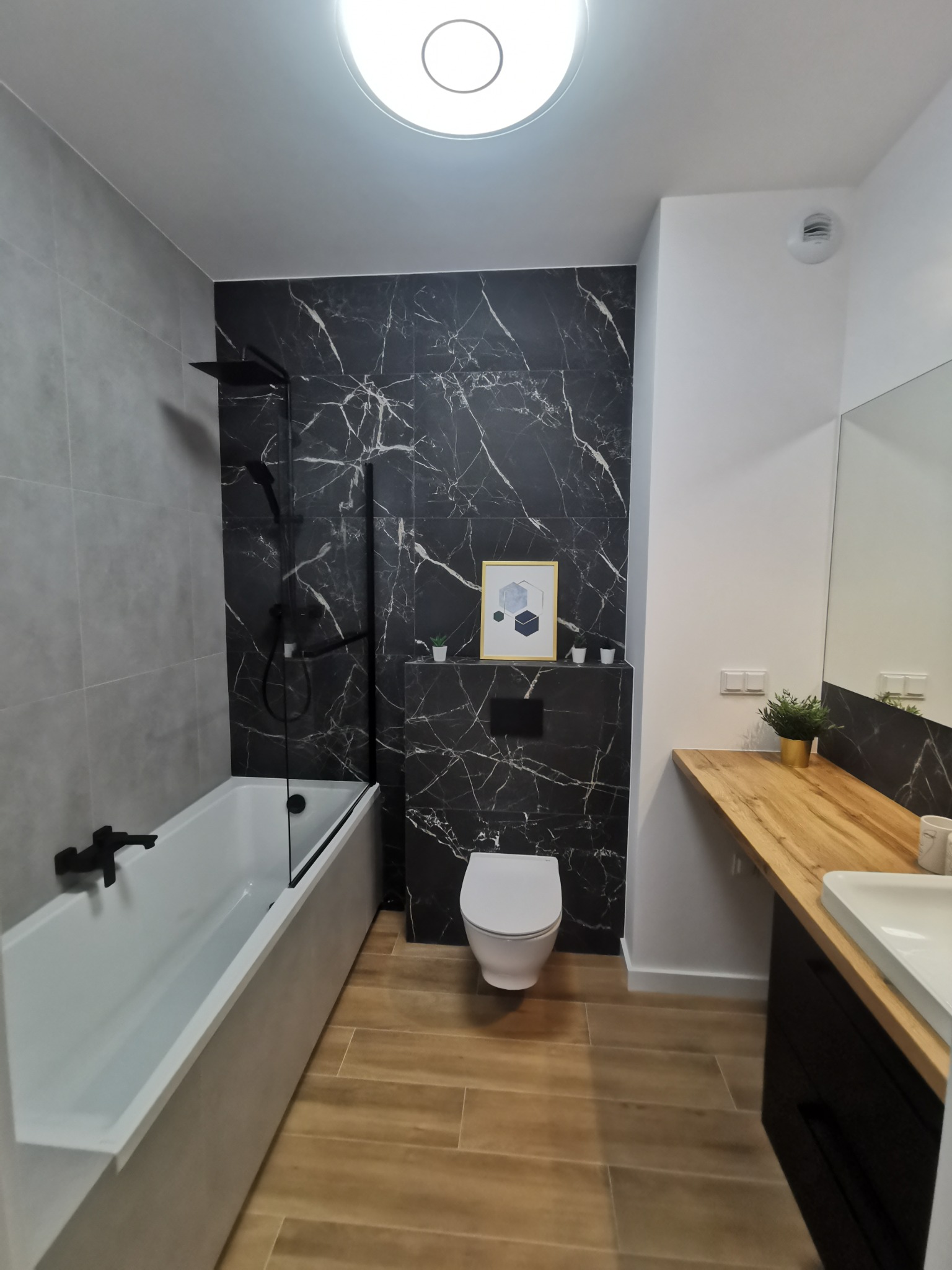 Nowoczesna łazienka z wanną z czarną armaturą, czarną ścianą z marmurowym wzorem, drewnianym blatem umywalki i toaletą podwieszaną, oświetlona okrągłą lampą sufitową.