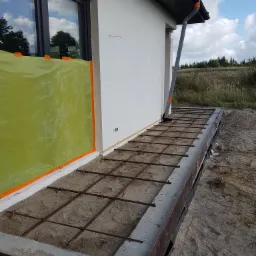 Przygotowanie pod wylewkę betonową tarasu przy budynku z zabezpieczonym oknem folią ochronną, widoczna siatka zbrojeniowa i szalunek.